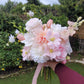 Bridal bouquet of pink and white flowers held by a person outdoors with greenery in the background