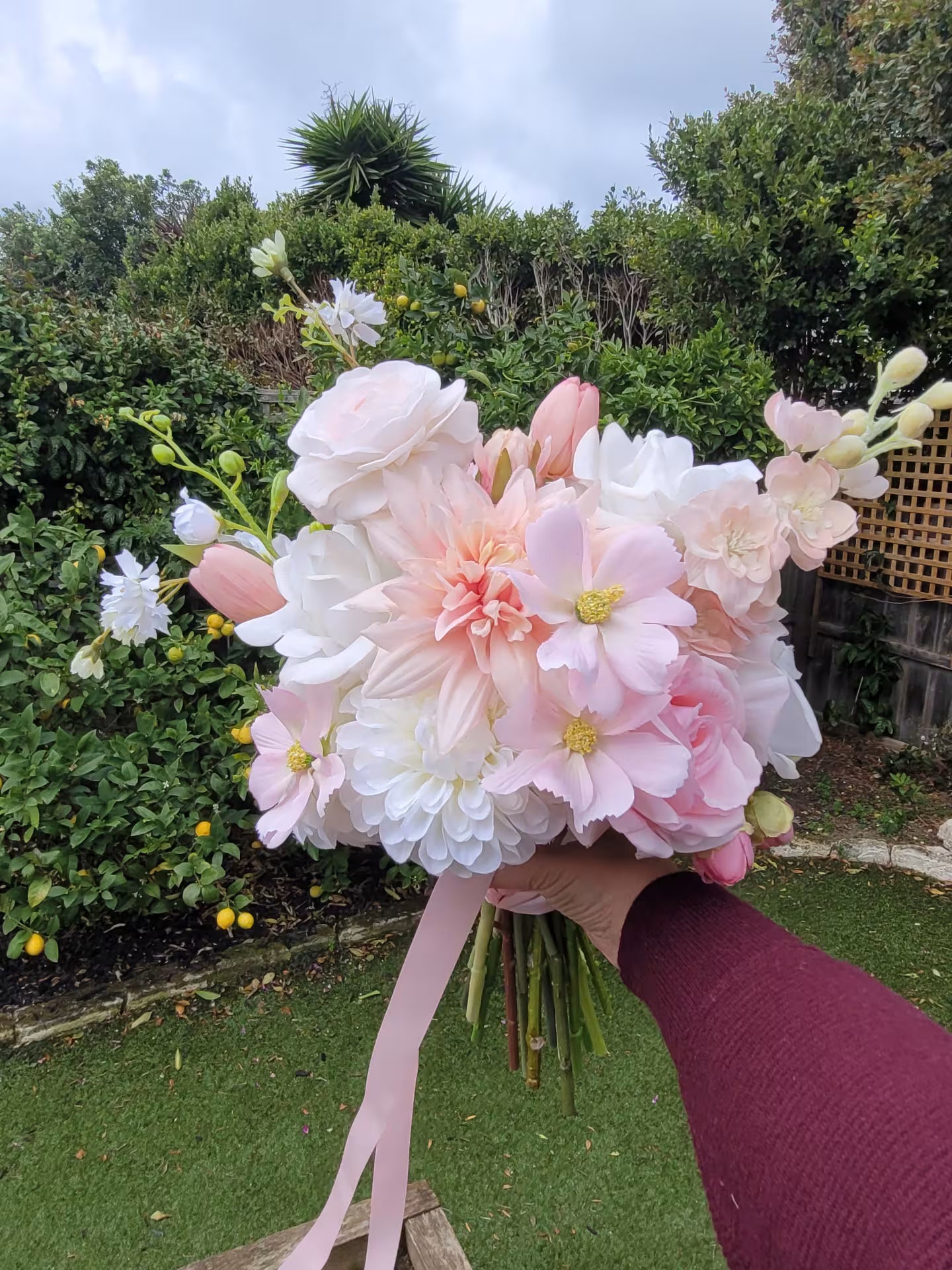 Bridal bouquet of pink and white flowers held by a person outdoors with greenery in the background