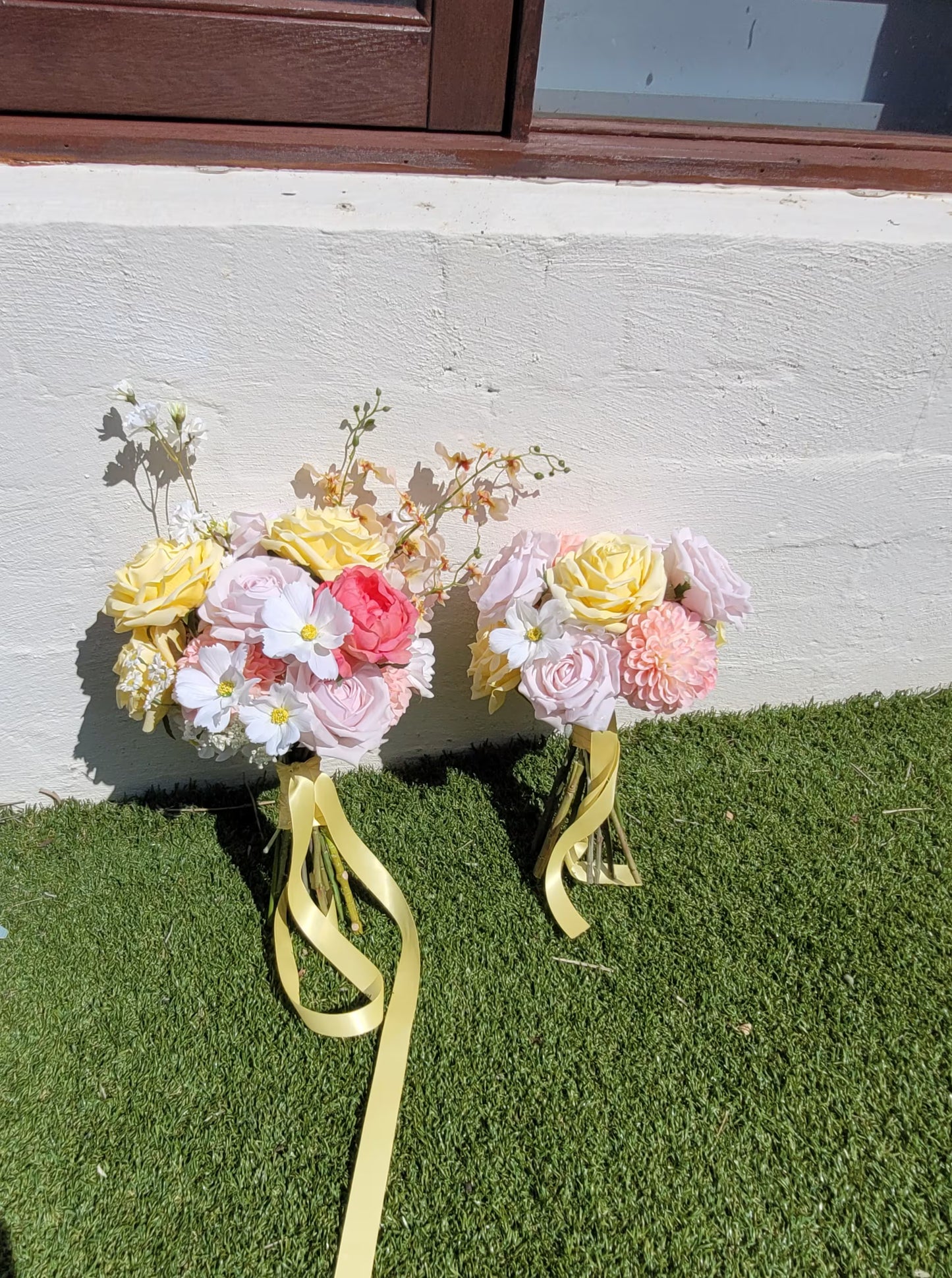 Two floral bouquets with yellow ribbons on a grassy surface against a white wall.