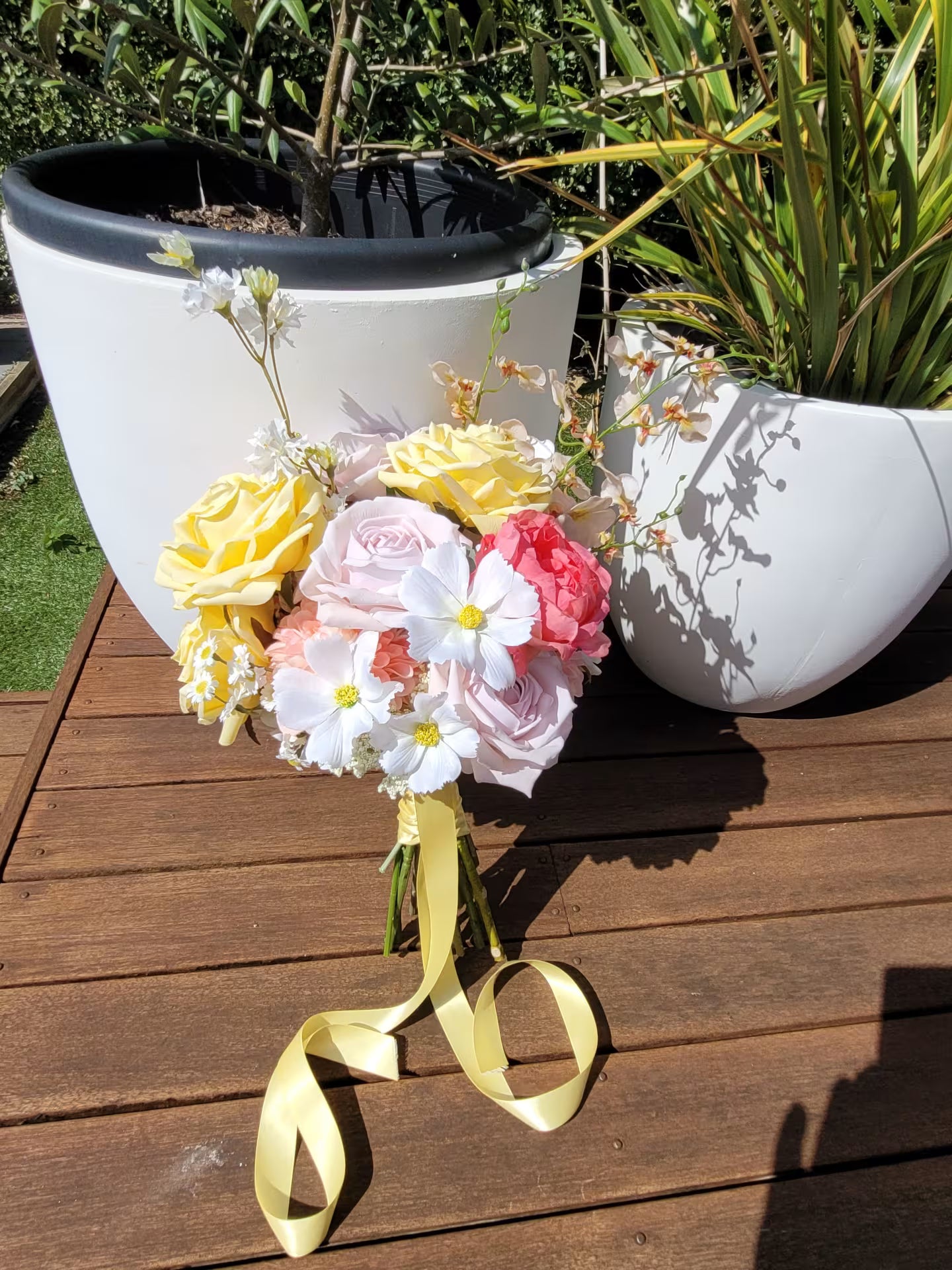 Bouquet of flowers with yellow, pink, and white colors on a wooden surface with plant pots in the background.