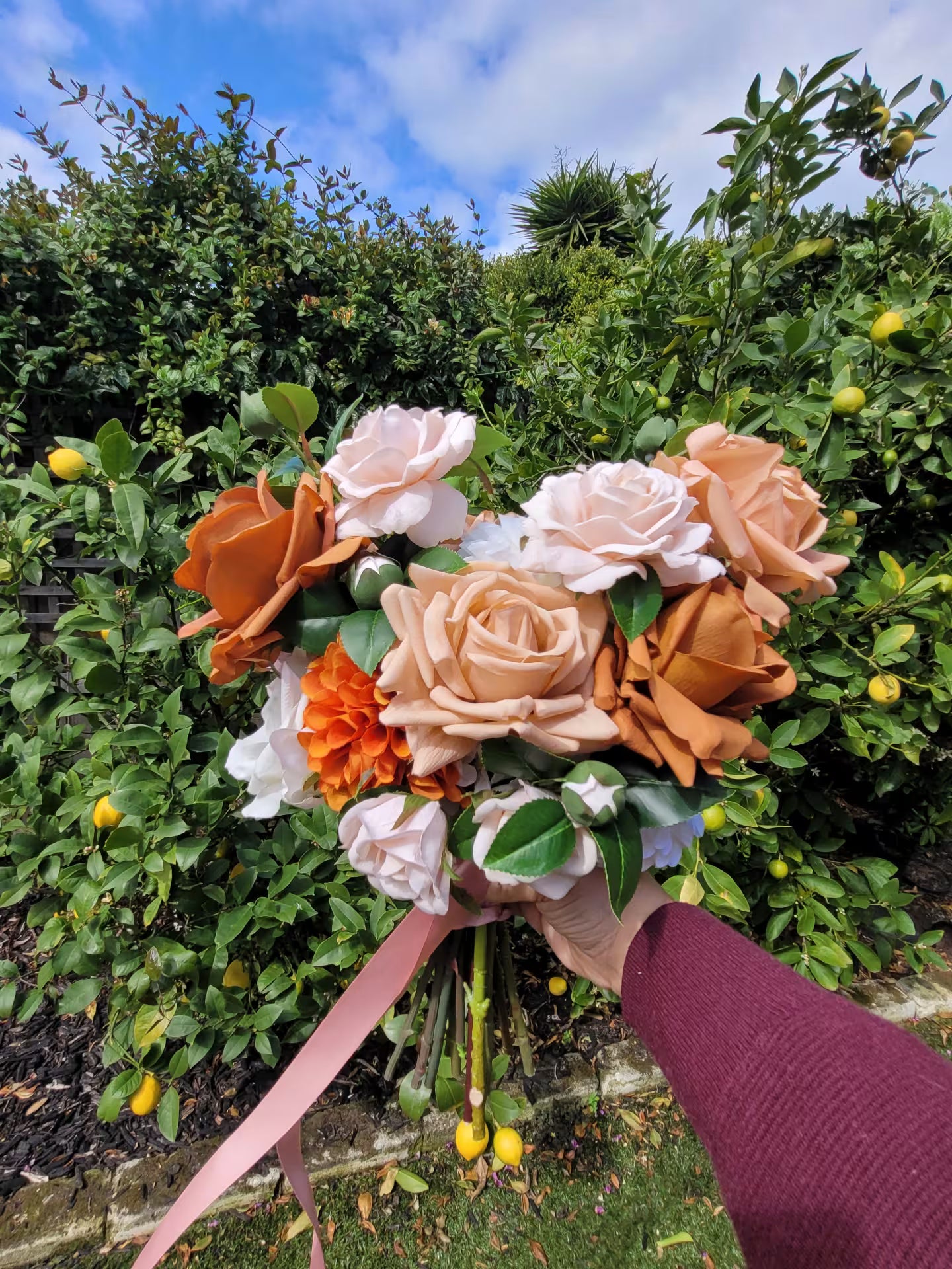 Bouquet of flowers held in front of a garden with greenery and blue sky.